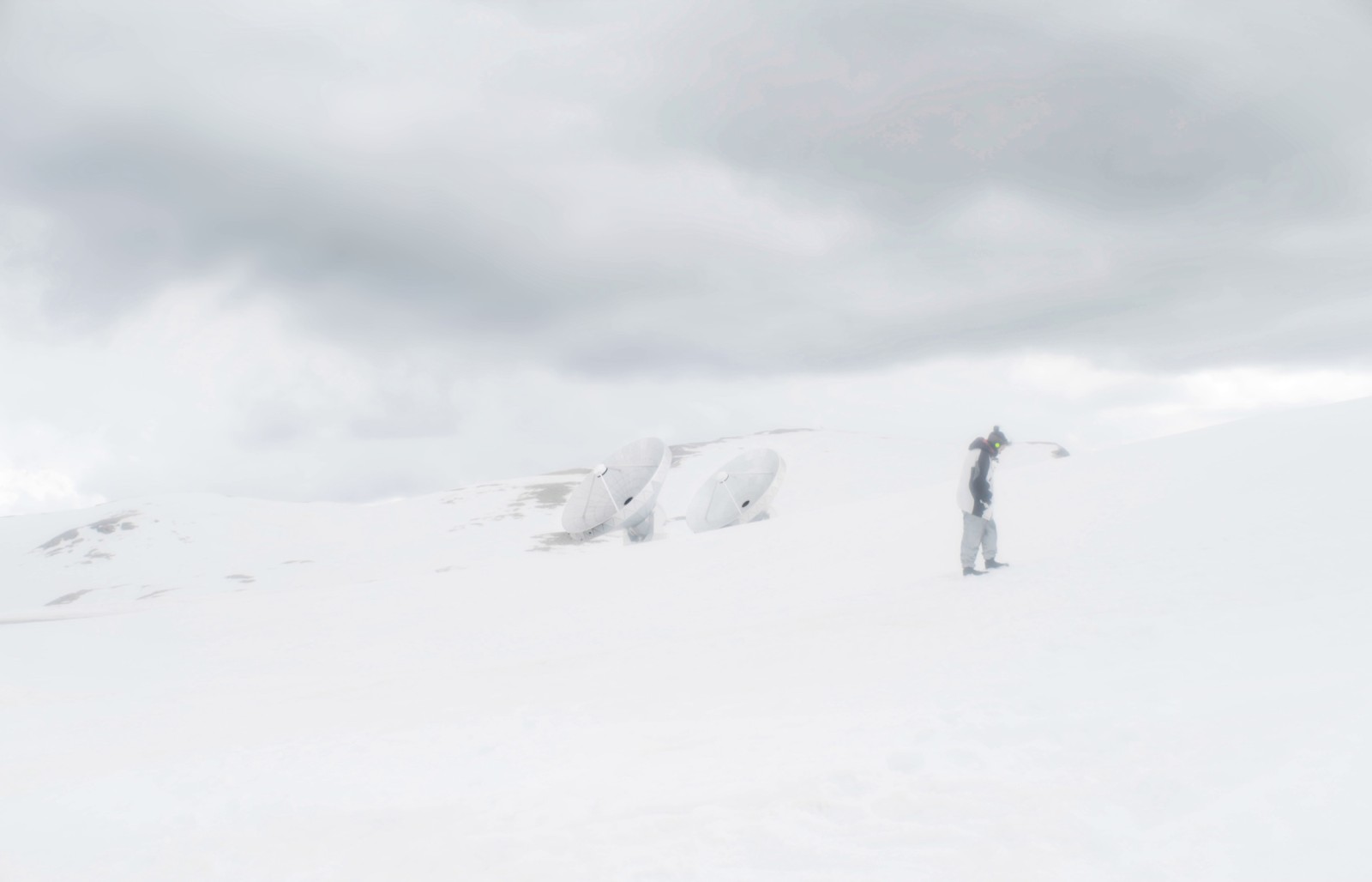 A film still of a wide shot on a snowy mountain top, we see somebody with headphones on strolling in the snow and two large radio telescopes in the distance.