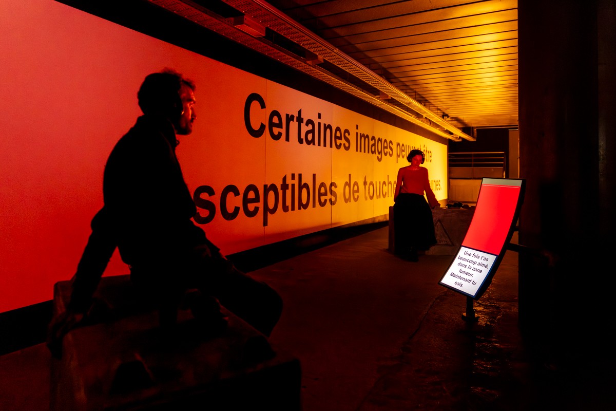 Dim red-lit underground space; a silhouetted figure sits in the foreground while a person in red stands by a long wall printed with French text, beside a glowing red screen.
