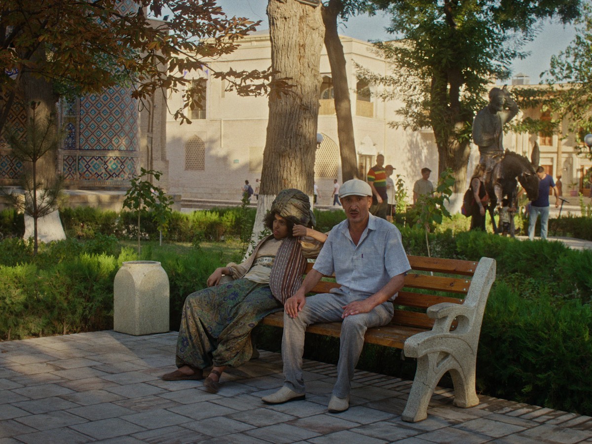 In a sunlit park framed by trees and patterned architecture, two people – a slim man in a short-sleeve short and with a white hat and a Nasreddin figure in tradditional wear – sit on a wooden bench in a quiet, contemplative pause while passersby and a statue of a man on top of a donkey animate the background.