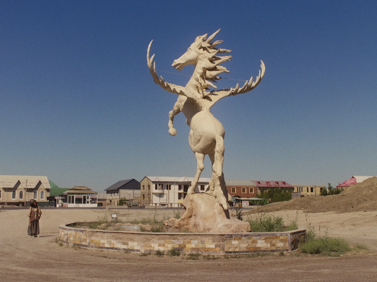 In a wide, sunlit square on the edge of a developing neighborhood, a solitary figure – Nasreddin – pauses beside a monumental sculpture of a mythical winged horse rearing atop a dry fountain. The vast blue sky and sparse surroundings lend the scene a surreal, dreamlike sense of isolation.