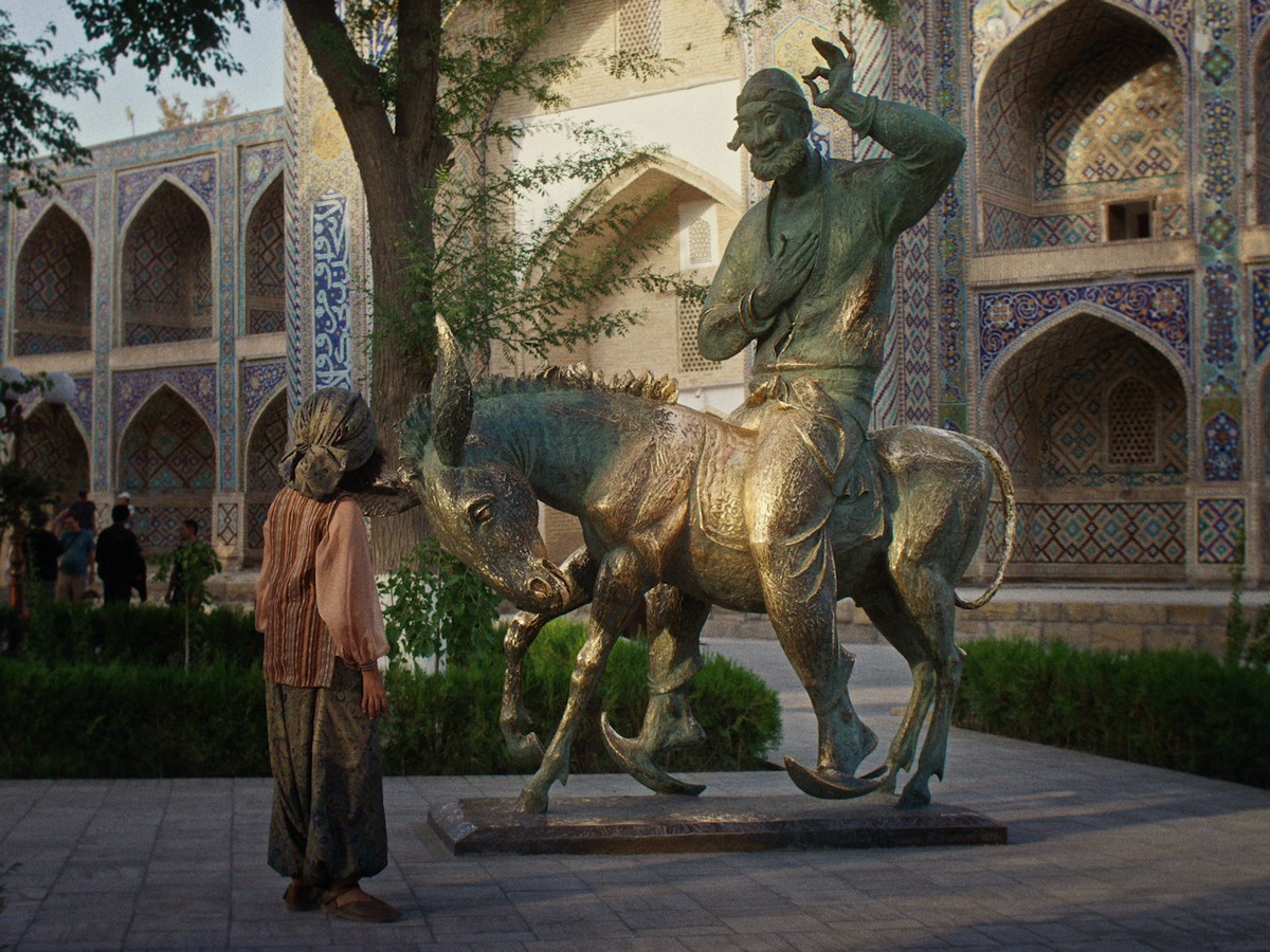 In a sunlit courtyard framed by intricate tiled arcades, a lone figure seemingly from the past stands before a bronze statue of a smiling rider on a donkey, it's Nasreddin. Dappled light from a nearby tree softens the scene, creating a reflective, almost surreal moment as the figure silently regards the statue.