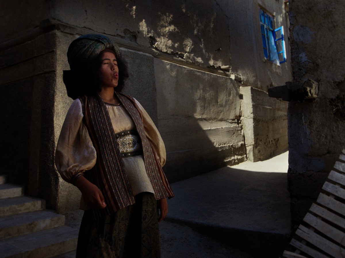 In a narrow earthen alley, a person in layered traditional dress leans against a weathered wall, eyes closed and lips pursed as if whistling. Warm sunlight and patches of shadow fall across the adobe buildings, while an open blue window draws the eye into the stillness of the scene.