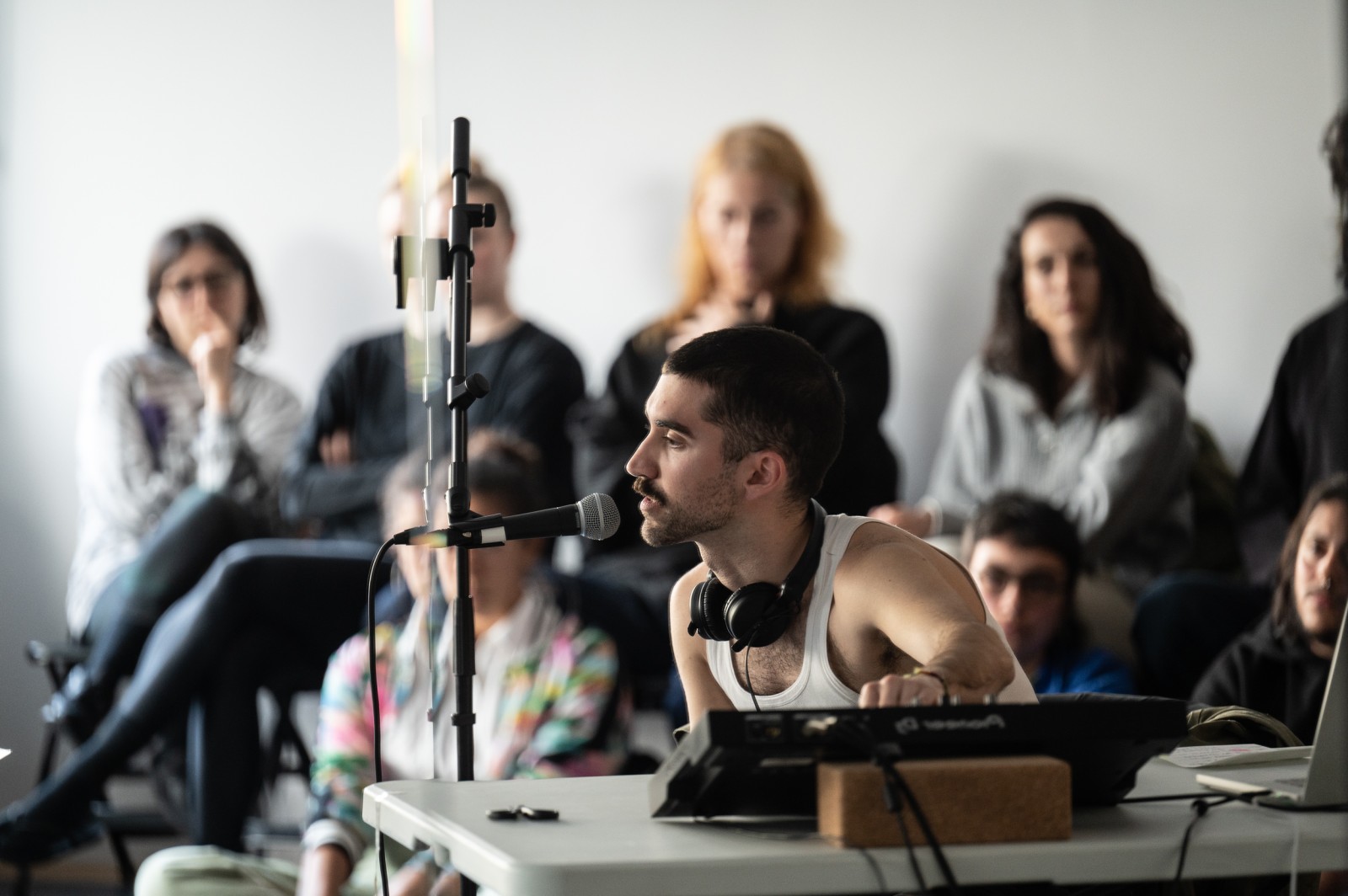 A male-presenting artist performs into a microphone while seated at a table with audio gear, DJ controller and several stands with an audience seated behind him in a studio space.