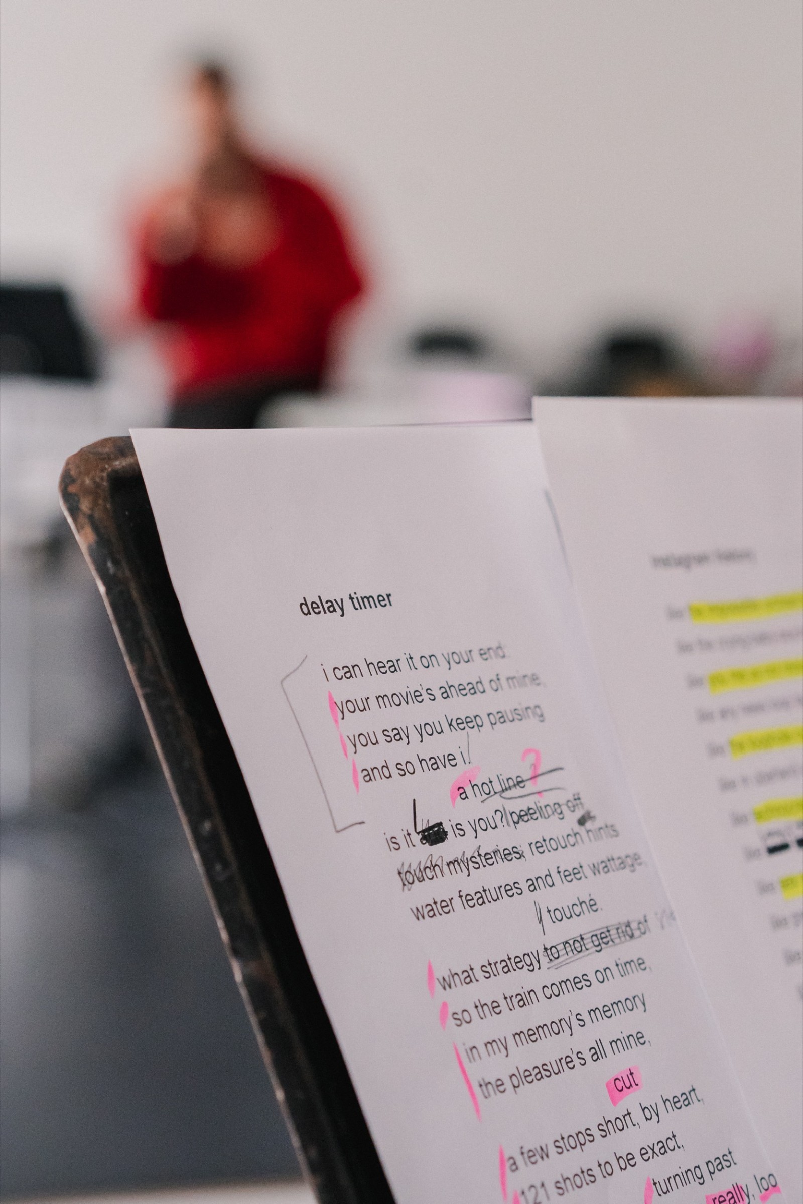 A detail shot of a poem called "delay timer" with various pen and marker annotations. In the blurry background we see a make artist in a red sweater, very blurry.