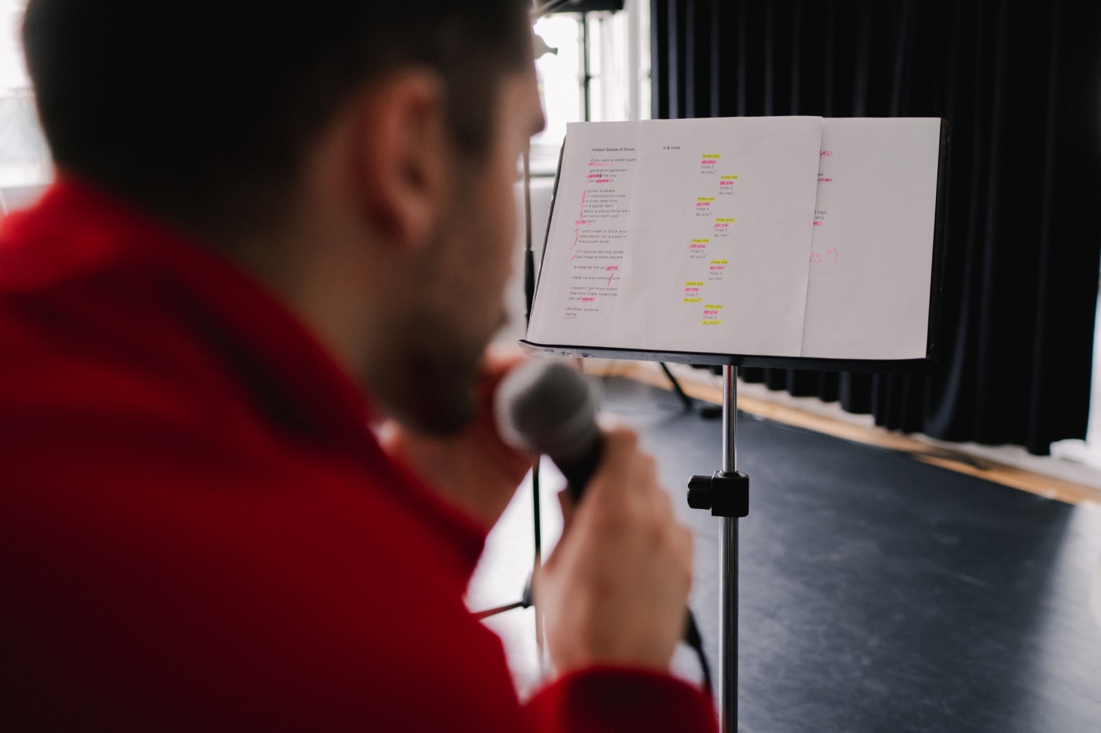 A male artist performs to a microphone, reading annotated poetry on a sheet music stand. An over-the-shoulder shot.