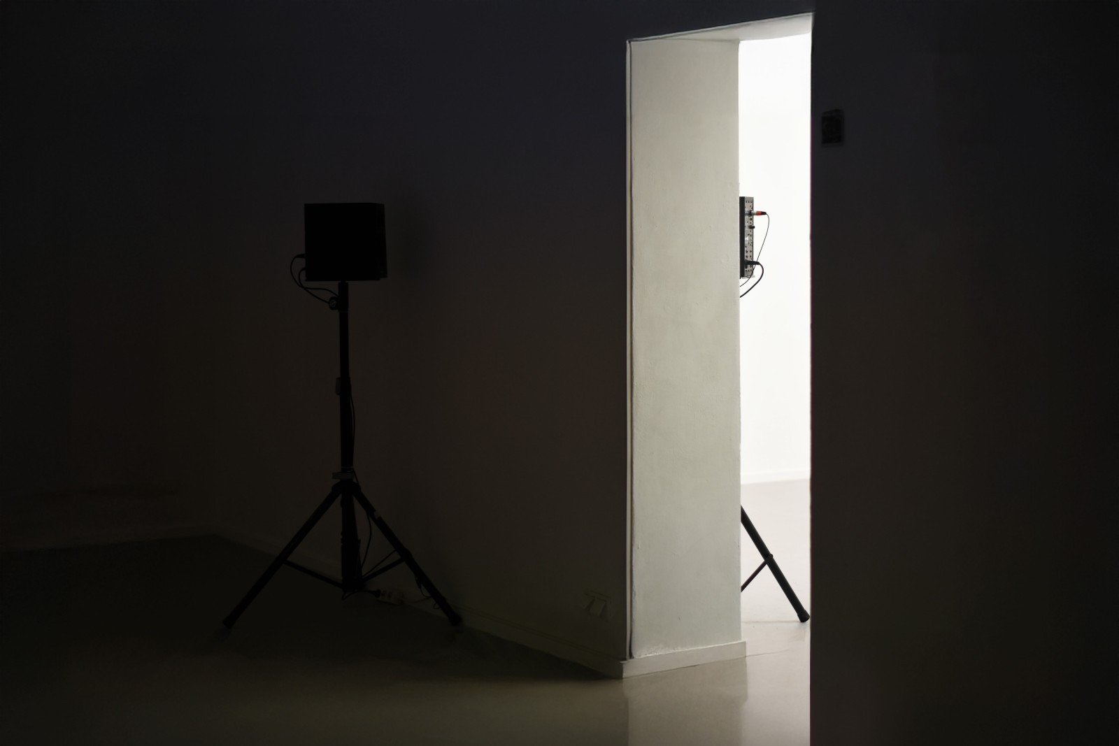 A dark room with a bright doorway revealing part of a white wall and a tripod-mounted device, with another tripod silhouette standing in the shadows.