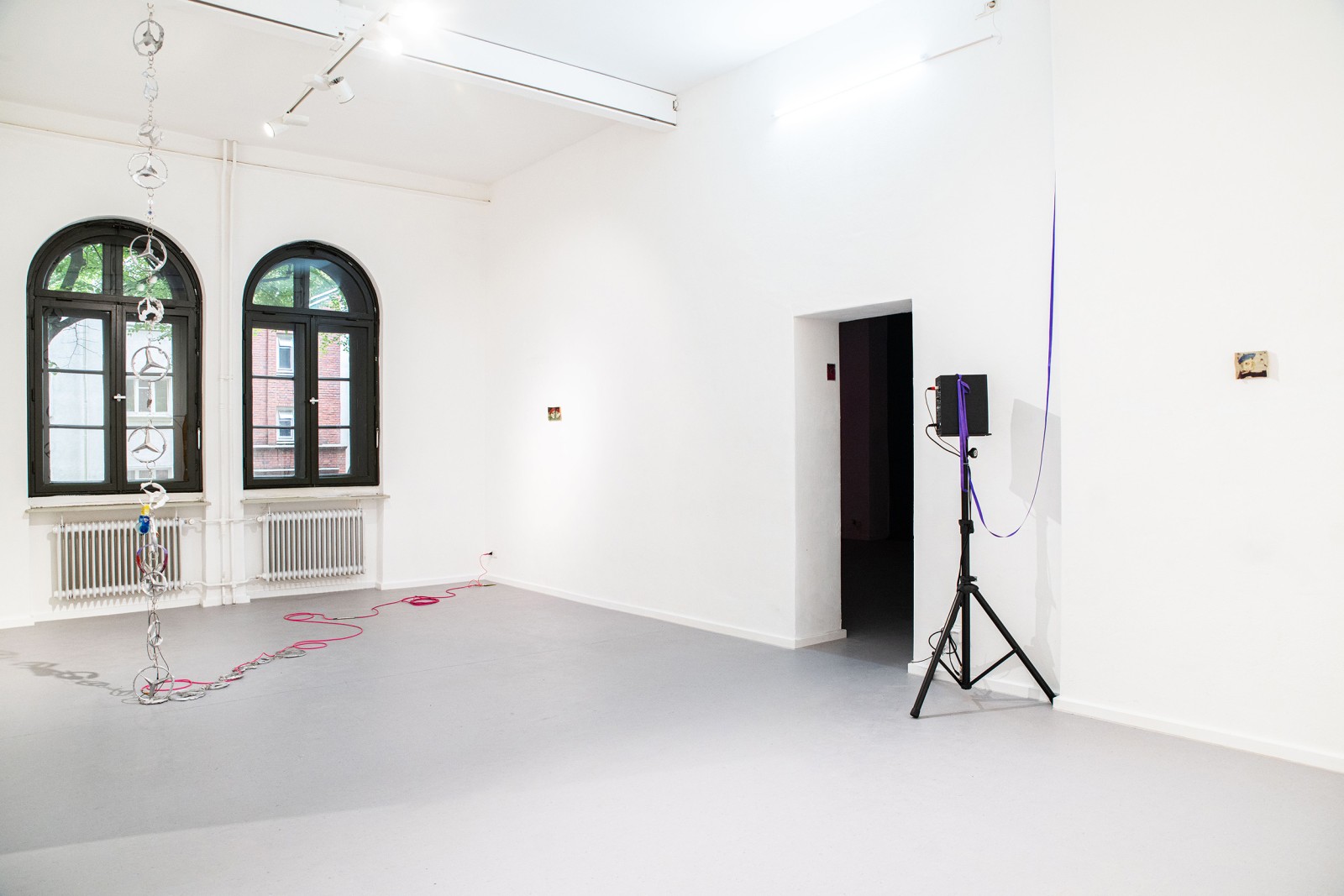 A bright white gallery room with arched windows, a hanging chain sculpture, a red cable on the floor, and a tripod-mounted device near a doorway.