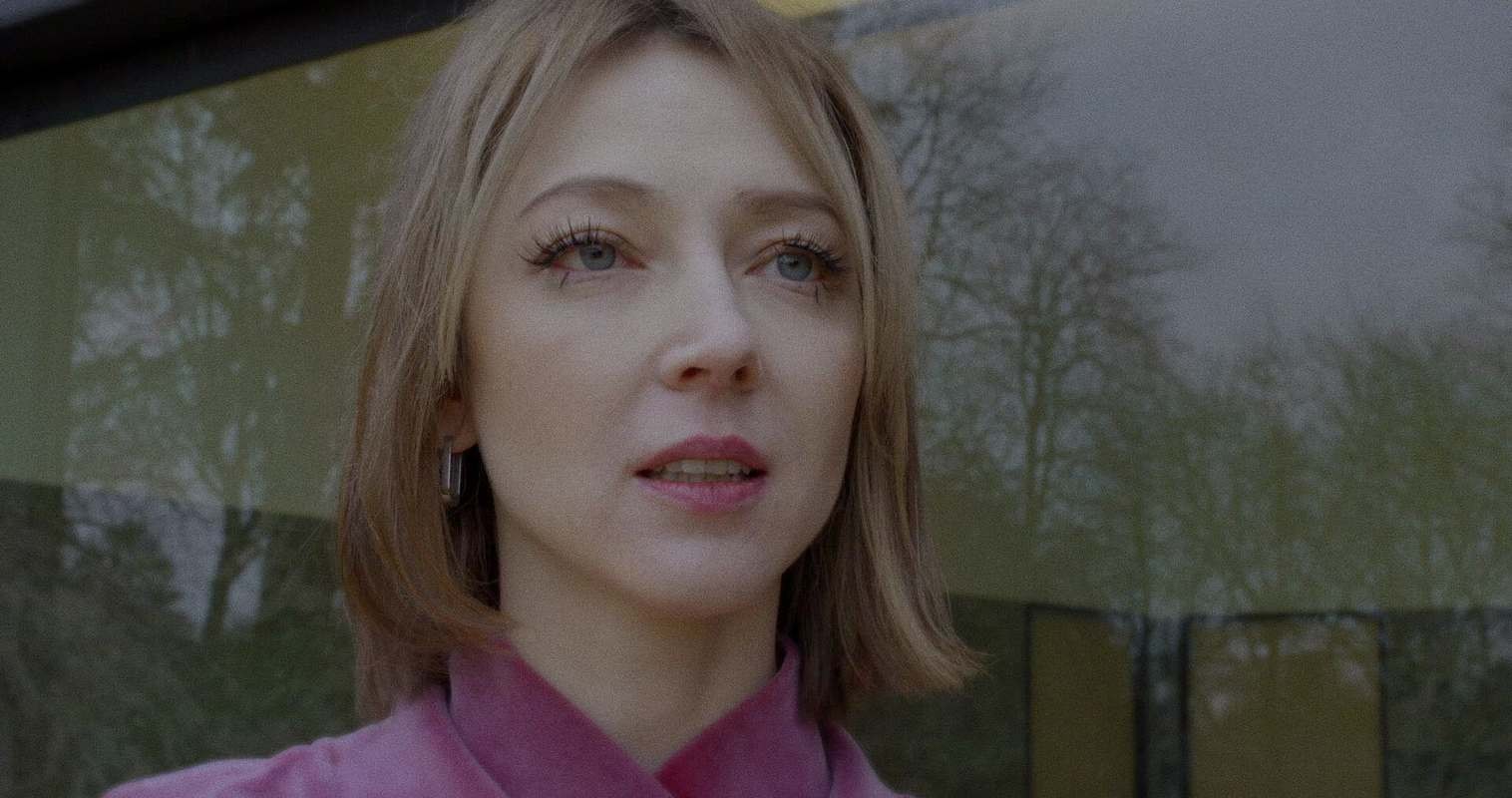 Close-up of a person with short light brown hair, with prominent lashes and blue eyes, wearing a pink sweater, standing outdoors in front of a reflective glass wall. They look slightly upward with parted lips, while bare trees are reflected in the glass behind them under overcast light.