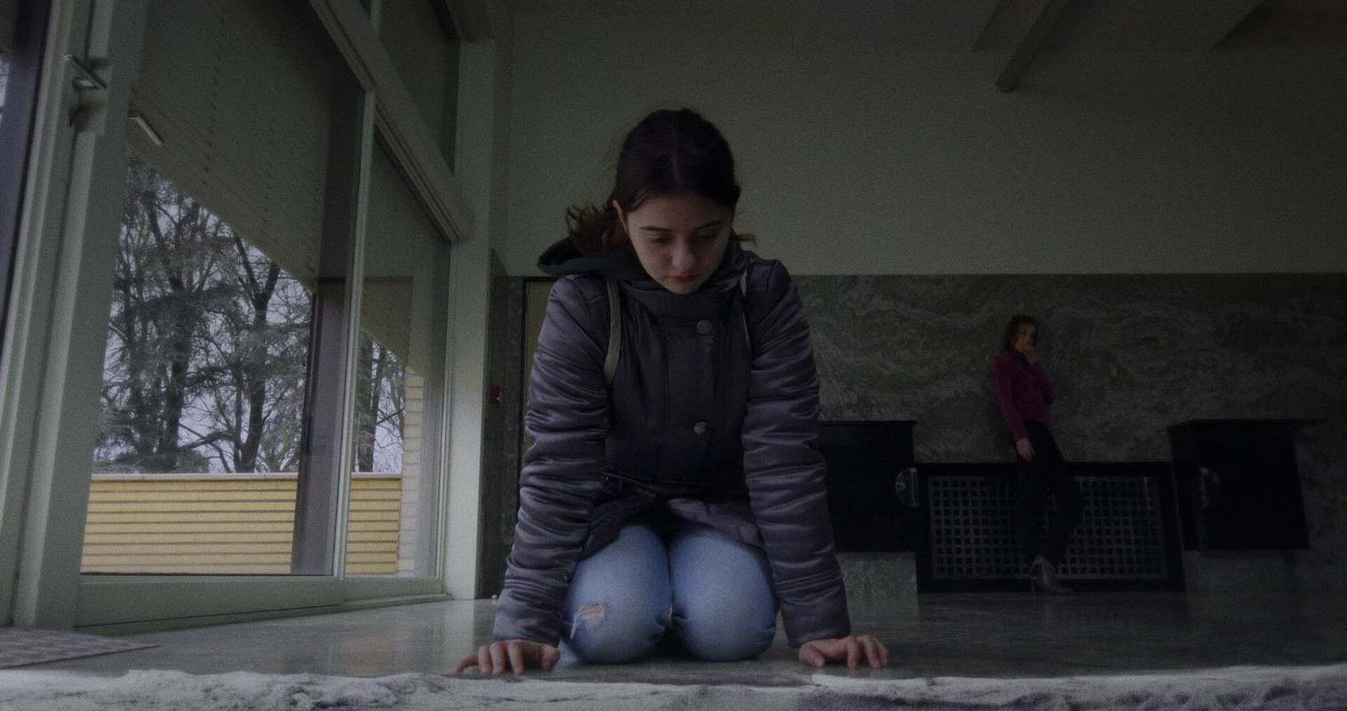 A young girl kneels on a tiled floor in a dimly lit, modern interior, looking down with their hands resting on the marble floor. Large windows on the left reveal bare trees outside, while another person in a pink sweater stands blurred in the background near dark fixtures.