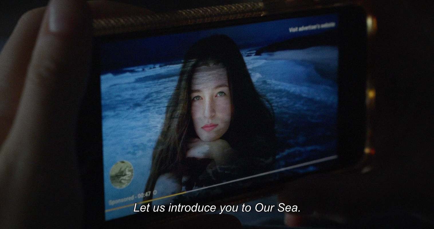 A hand holds a smartphone displaying a video advert (with the usual video progress bar and associated info) of a young person with long dark hair against a blue ocean backdrop. The subtitles read "Let us introduce you to Our Sea."