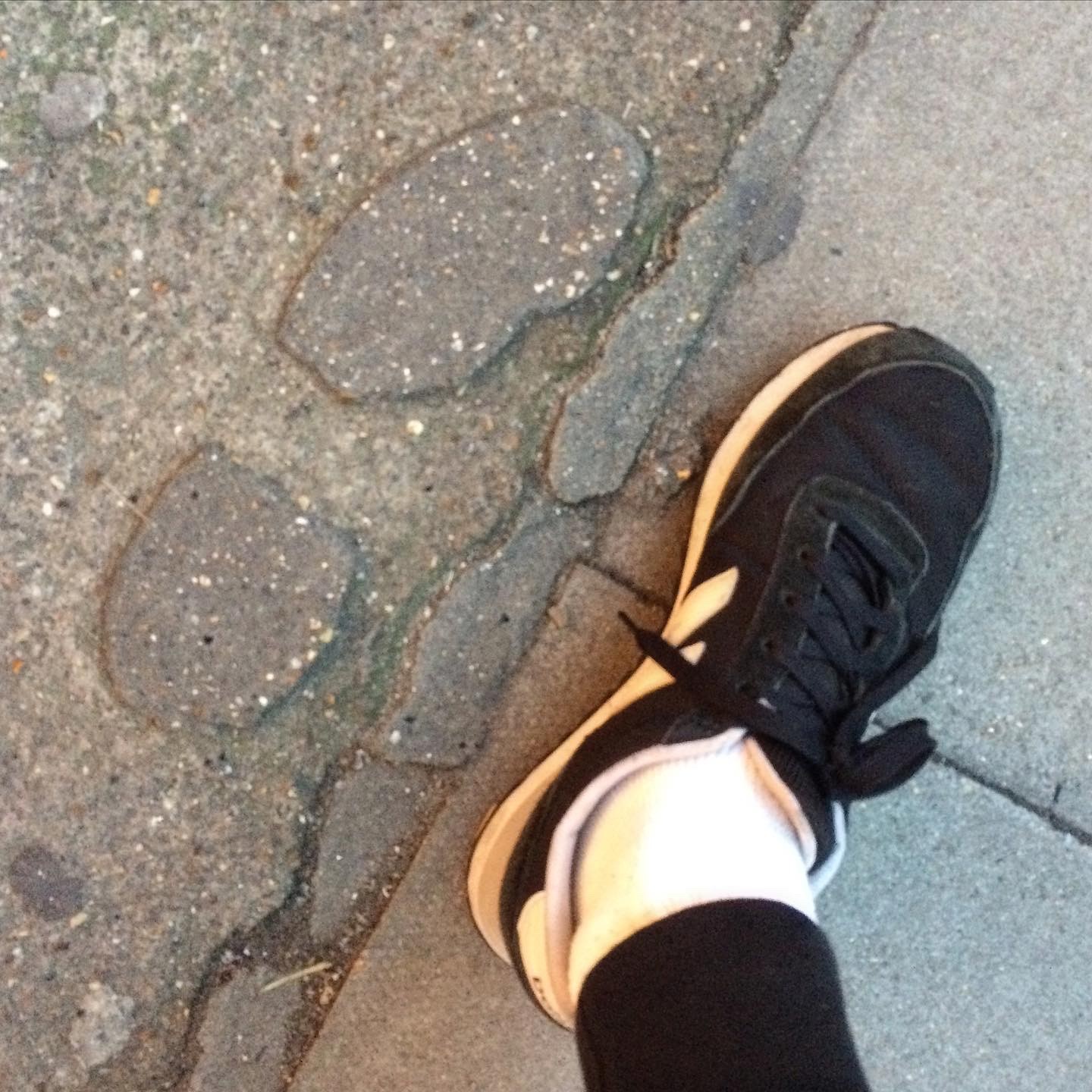 A black sneaker on the sidewalk, on the right. On the left, a footprint on the concrete from a similar shoe size.